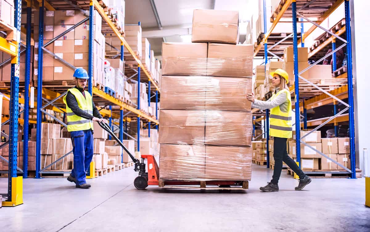 A man pulls an oversized pallet on a pallet truck in a warehouse while a woman supports the pallet contents.